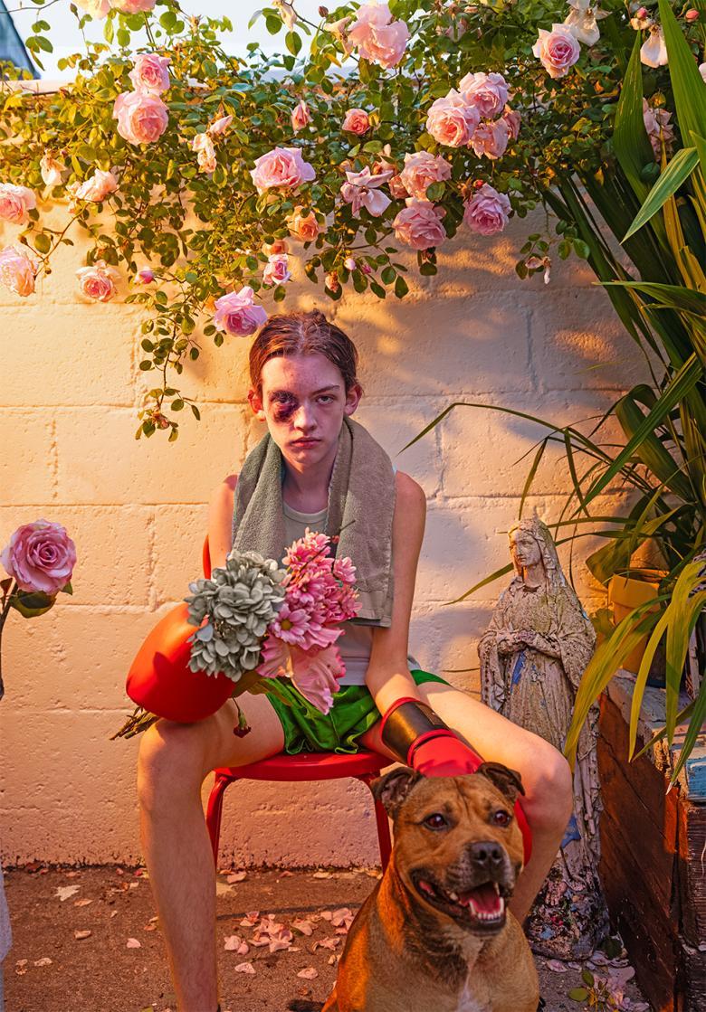 A male figure sits in a garden surrounded by rose plants. He has a black eye, is wearing boxing gloves and holding flowers and holds the lead of a dog.