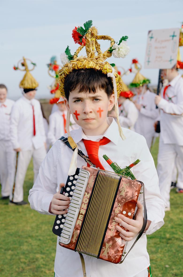 A young boy wears a traditional 'Biddy Boy' outfit and holds an accordion.