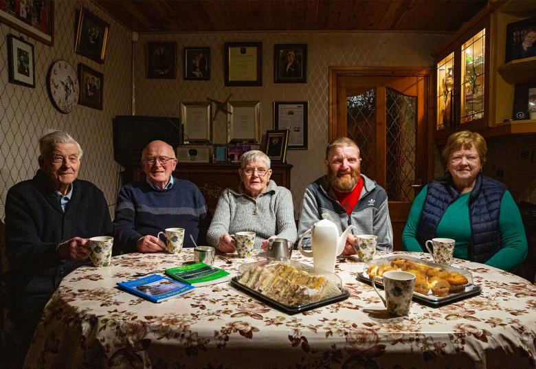 A photograph of three men and two women, sitting behind a table covered in cups of tea and sandwiches.