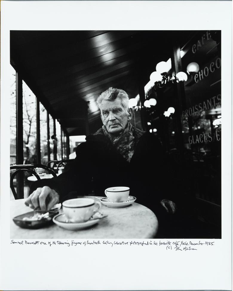 Black and white image of an older male figure sitting at a Parisian cafe, with two coffee cups on the table in front of him.
