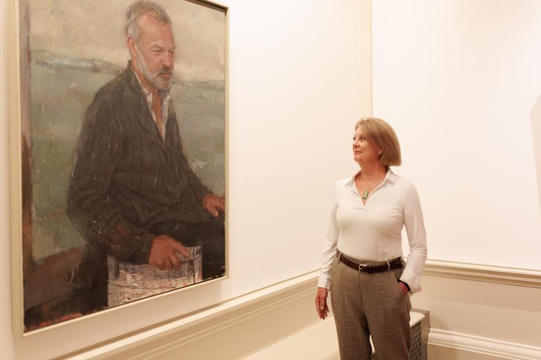 A woman stands with one hand in her pocket looking at a portrait hanging beside her, of a man seated. 
