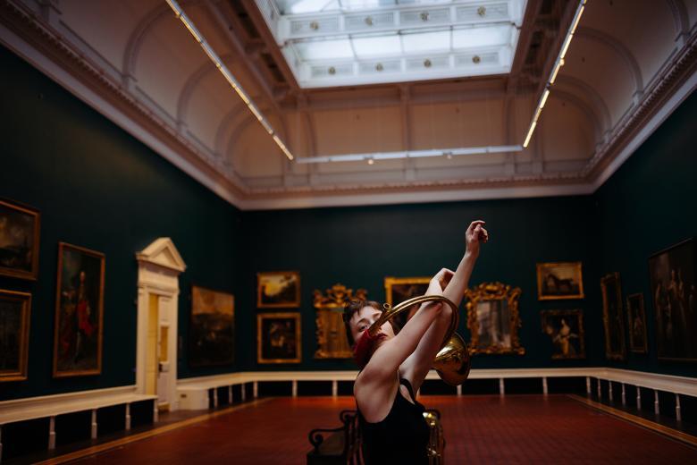 A figure stands in a gallery filled with paintings, arms outstretched through two metal rings.