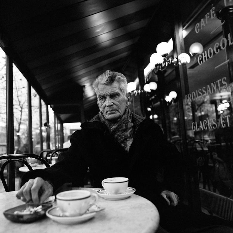 Black and white photograph of Samuel Beckett in later years, sitting at a marble table outside a cafe in Paris with two teacups on the table.