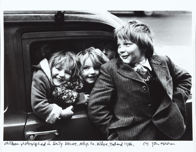 Black and white photograph of three young children laughing in front of a car.
