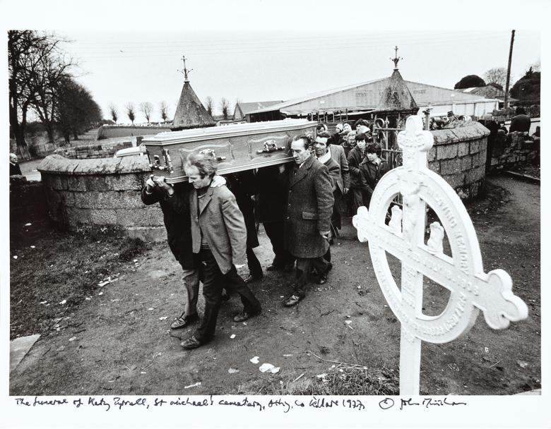 Black and white photograph of pallbearers carrying a coffin in a graveyard, followed by a crowd of mourners.