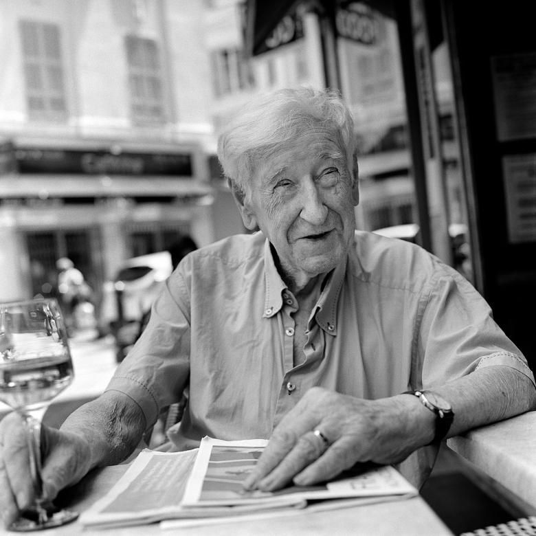 Black and white photograph of an older man with white hair and a short sleeved shirt, sitting at a table in front of a cafe in Nice.