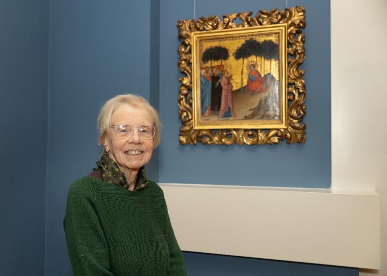 A woman in a green jumper stands smiling beside a painting in an intricate gold frame.