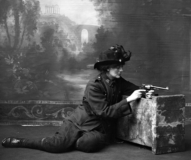 La condesa Constance Markievicz en uniforme, arrodillada contra un accesorio de estudio con una pistola, estudio.