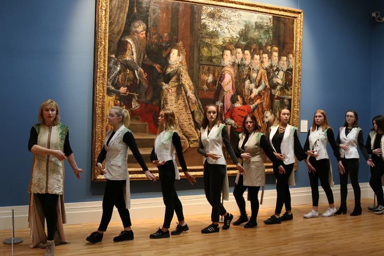 A group of nine women wearing cream jackets walk in sequence through a Gallery, in front of a large painting