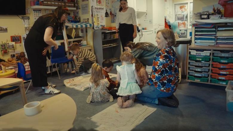 A creche with young children and three adults, playing amongst a toy shopfront, blocks and colouring materials