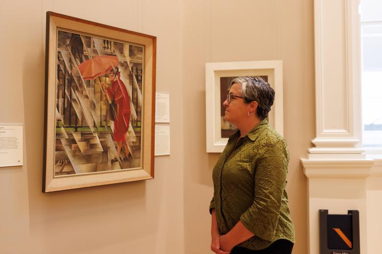 A woman in a green shirt stands in a gallery looking at a painting on the wall of a woman in red with an umbrella over her head