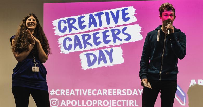 Two people stand on stage in front of a bright pink screen with the words Creative Careers Day written across it.