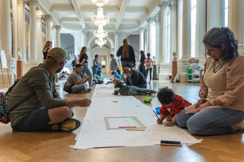 A group of adults and children sit on the floor of a large hall, drawing on a long sheet of paper running the length of the room.