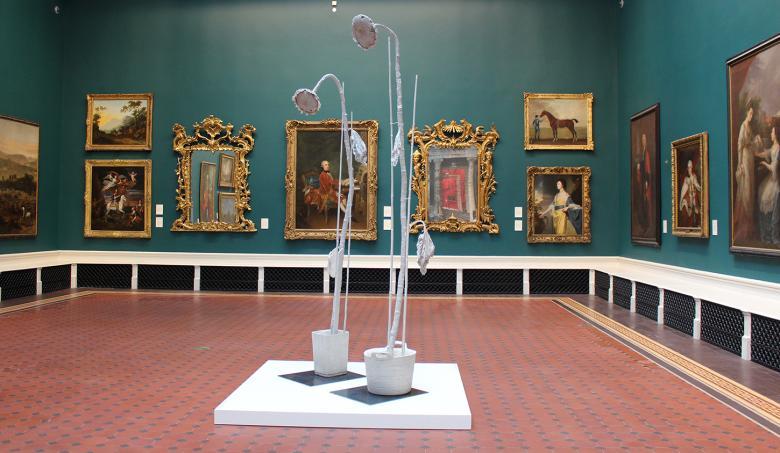 Two tall and spindly sculptures of sunflowers displayed on a low plinth in a gallery hung with gilt-framed oil paintings
