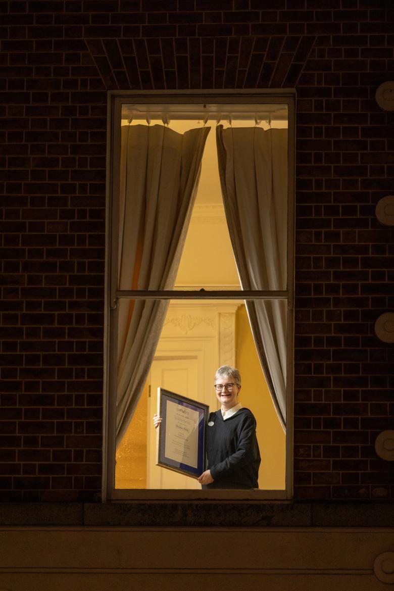 A figure stands at a window, holding a framed certificate.
