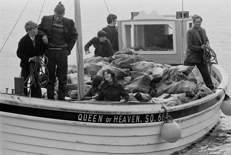 A black and white photograph of a boat, with seven passengers, some of whom are looking at the camera.