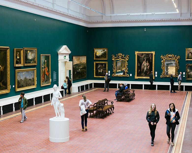 Photo of people walking through a green gallery room with gilt-framed paintings covering the walls and a white marble statue standing in the middle of the room.