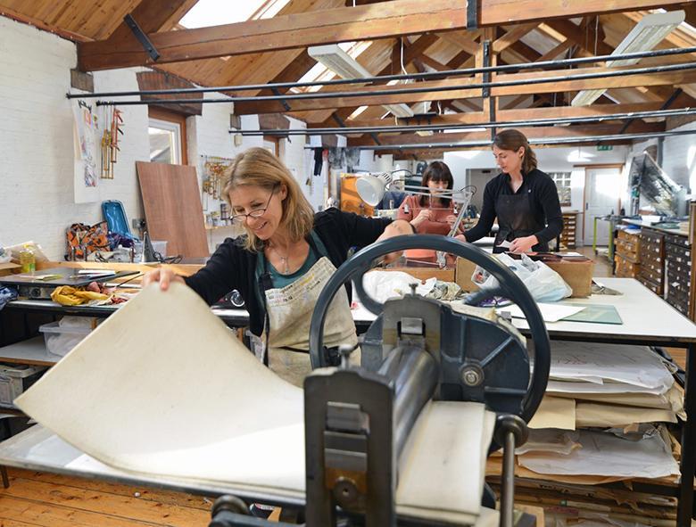 Colour photograph of a printmaking studio with one woman in the foreground running paper through a printing press.