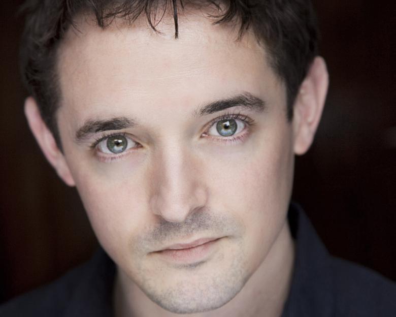 Closely cropped headshot of a man with dark hair, pale eyes and faint stubble.