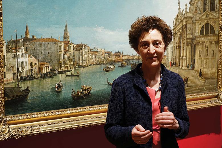 A photograph of Lucy Whitaker, Senior Curator of Paintings, Royal Collection Trust, standing in front of a painting by Canaletto.