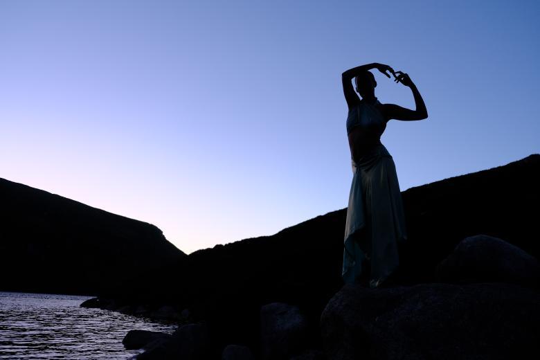 A dancer stands on the banks of a river with her arms linked above her head, silhouetted against a blue sunrise sky. 