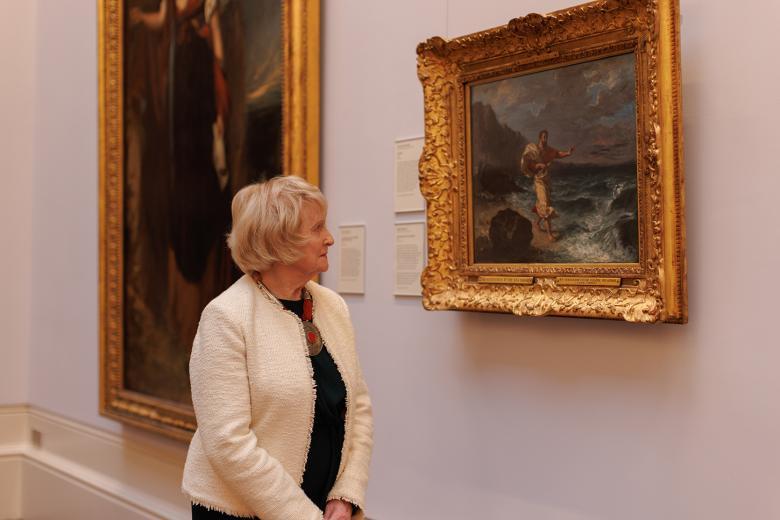 A woman in a white jacket and black dress stands in a gallery looking towards one of the paintings on the wall. 