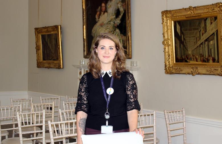 Sarah stands looking at the camera, resting her hand on a chair in front of her. Behind are chairs set out for a wedding, and hanging on the walls are three paintings in gold frames.
