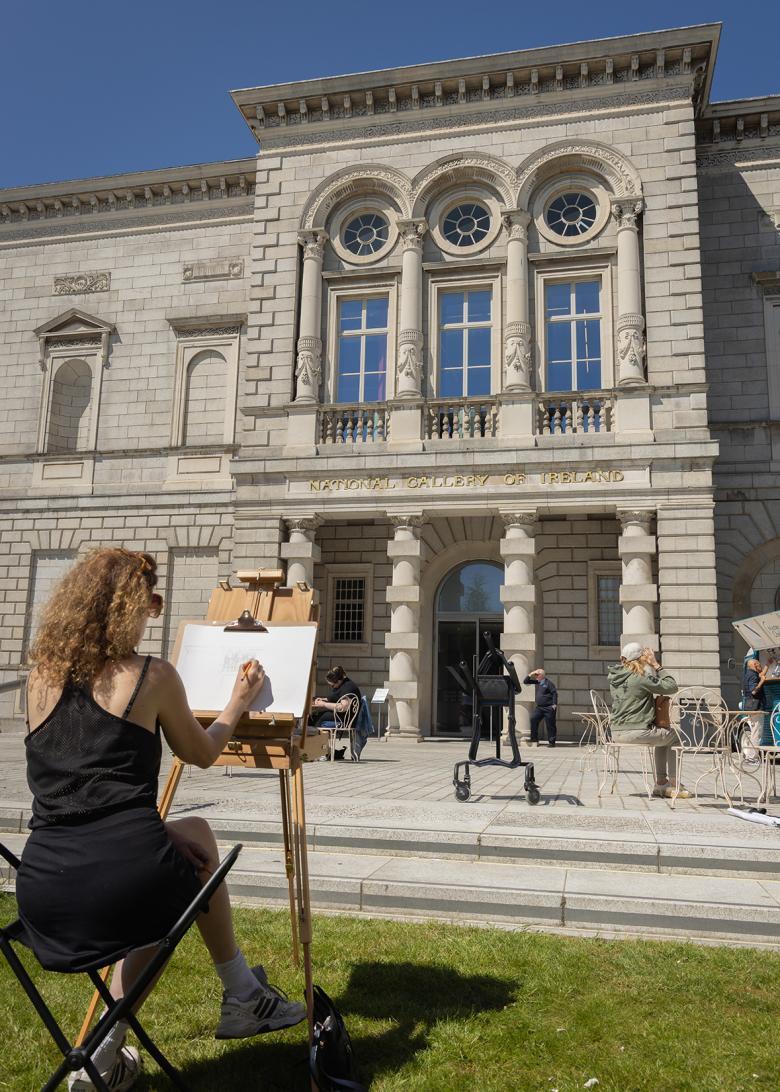 A woman sits and draws at an easel on the lawn in front of the National Gallery of Ireland