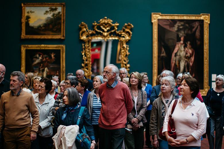 Photo of a group of people on a guided tour in a gallery, with gilt-framed pictures and mirrors on a wall behind them.