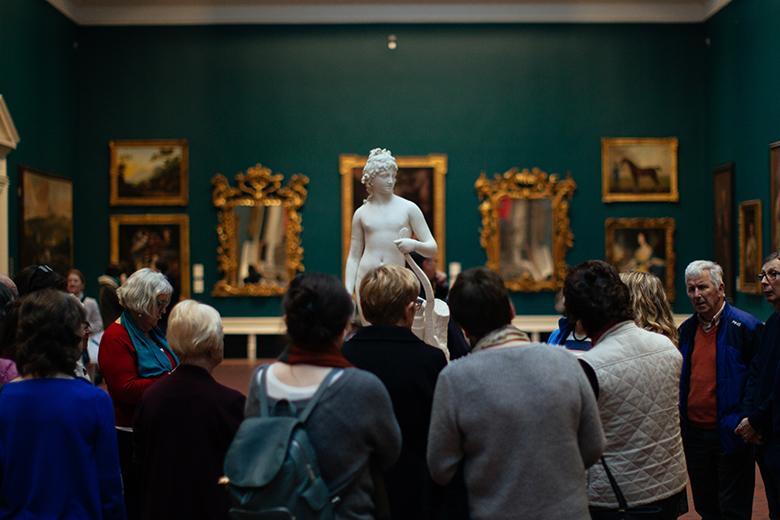 Photo of a group of people looking at a marble statue in a gallery room painted green and hung with gilt-framed paintings.