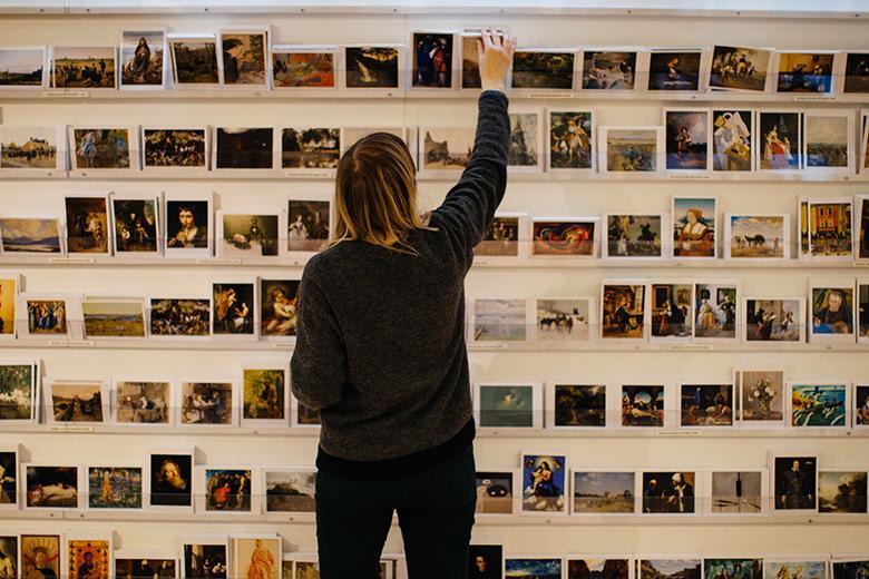 Photo of a woman standing, with her back to the camera, in front of a wall displaying postcards.