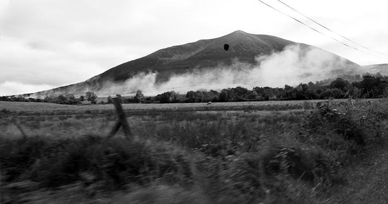 Black and white still from a video, shot from a moving car, showing countryside with a mountain in the distance shrouded in white smoke.