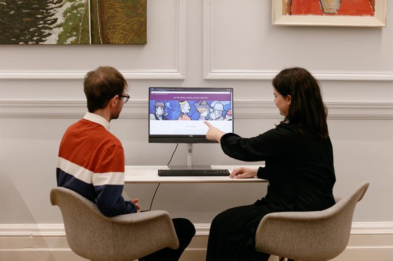 A man and a woman sit in front of a computer. The woman is pointing at an image on the screen.
