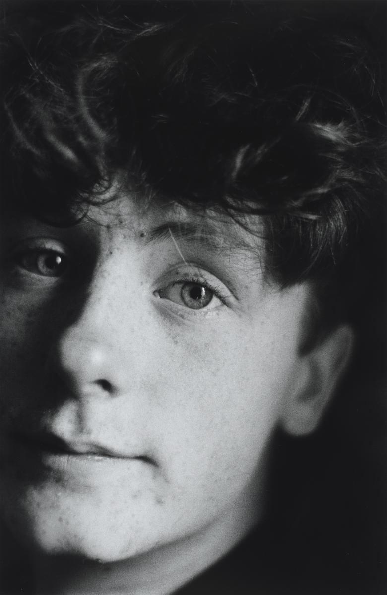 A black and white photograph of a young man's head. His face and curly hair fill most of the frame, and one side of his face is slightly out of frames looking directly at the camera and 