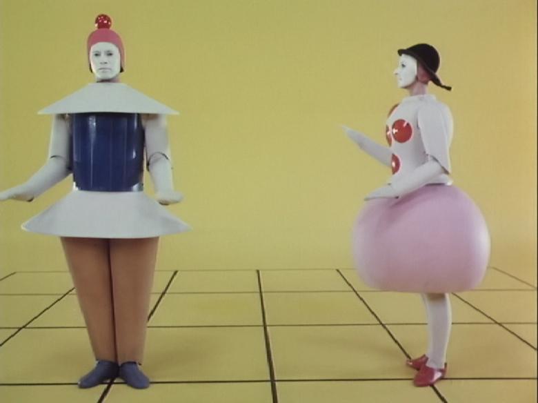 Film footage still of two figures wearing geometric costumes standing in a yellow space.