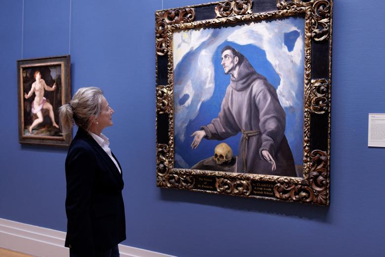 A woman in a black jacket stands gazing at a painting of Saint Francis on a blue Gallery wall