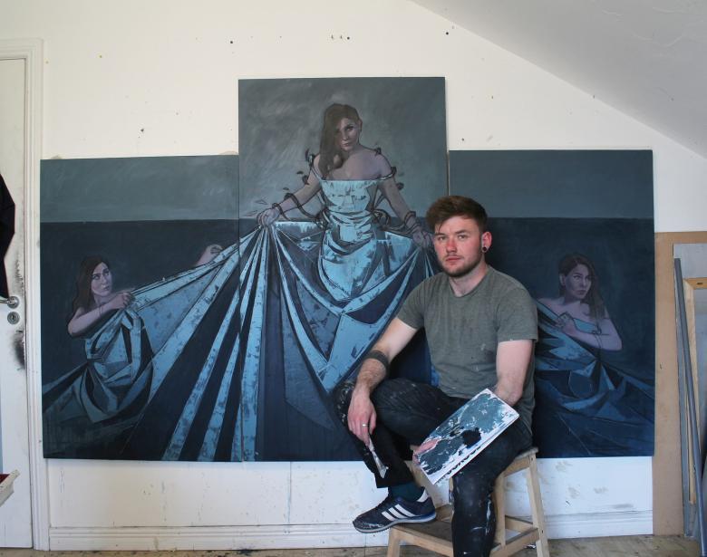 Photo of a male artist sitting on a stool, holding a paletter and paintbrush, in front of a large triptych portrait of three women.