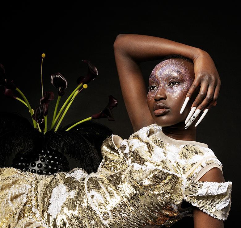 Photograph of woman with nail art, reclining beside plant, against black background.