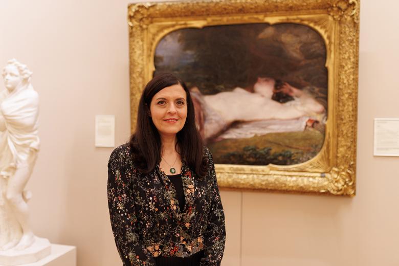A woman with long dark hair stands in front of a painting in an elaborate gilt frame.