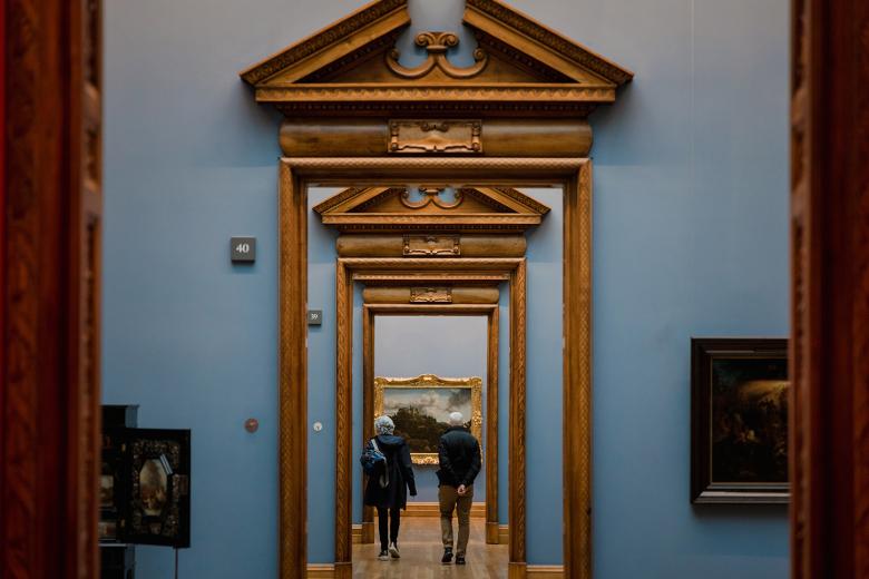 A photograph of a suite of galleries, looking through a series of ornate wooden doorframes. The walls are painted blue, and we can see some paintings hanging. In the distance, some people are walking through the rooms.