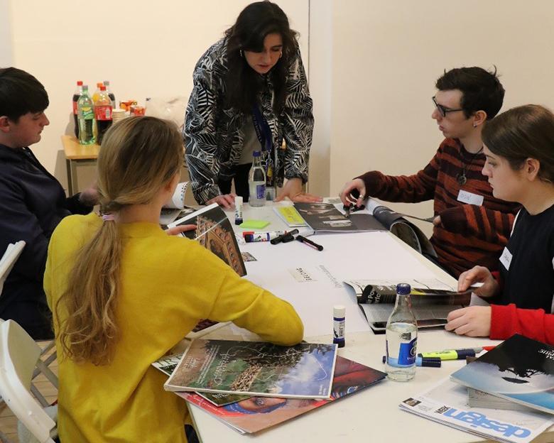 Photo of a group of young people seated at a table covered in magazines and art materials.
