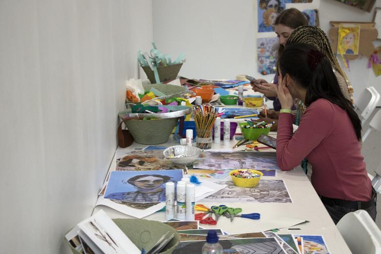 Two people sit at a long table covered in art supplies and materials