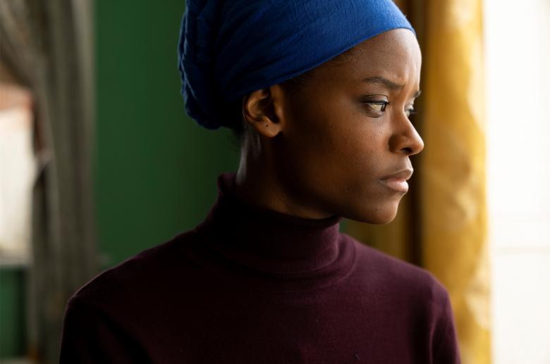 A film still of a young black female figure wearing a burgundy polo neck and a blue headscarf 