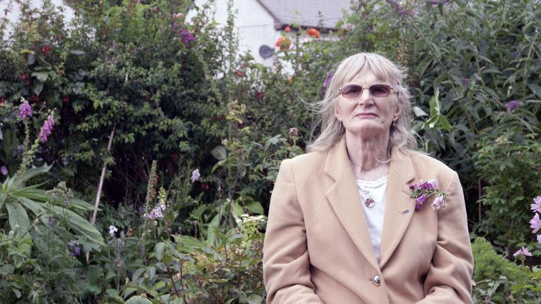 A woman in a beige coloured jacket stands in a garden, wearing dark glasses. On her lapel there is a foxglove; the garden behind is also filled with foxgloves.