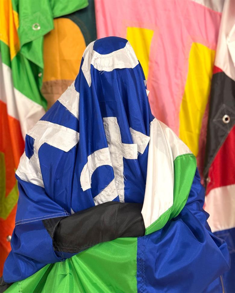 A figure is sitting with their arms folded covered in a large blue and green flag with white writing