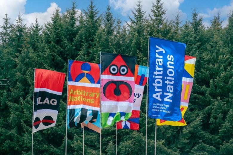 A group of colourful flags, flying in a strong wind, against a backdrop of spruce trees.
