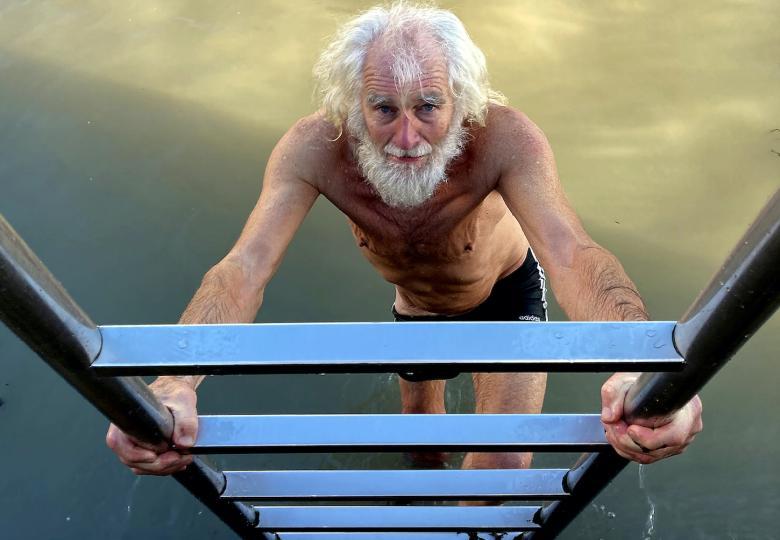 Photograph of an older male figure with long white hair and a beard wearing shorts climbing a ladder out of water