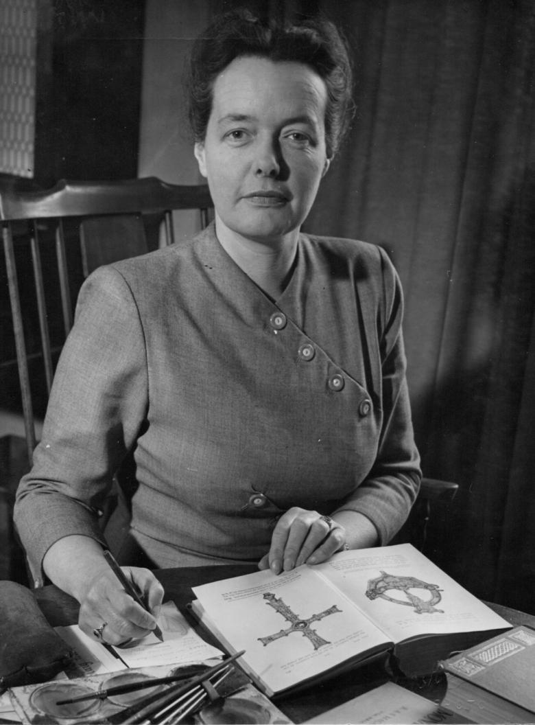 Photograph of Bea Orpen, sitting on a chair, looking at the camera with a book of her artwork open in front of her.
