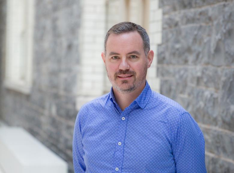 A photograph of a man in a blue shirt standing against a stone background