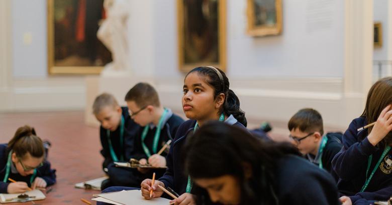 A group of young students sit in the Gallery working on clipboards